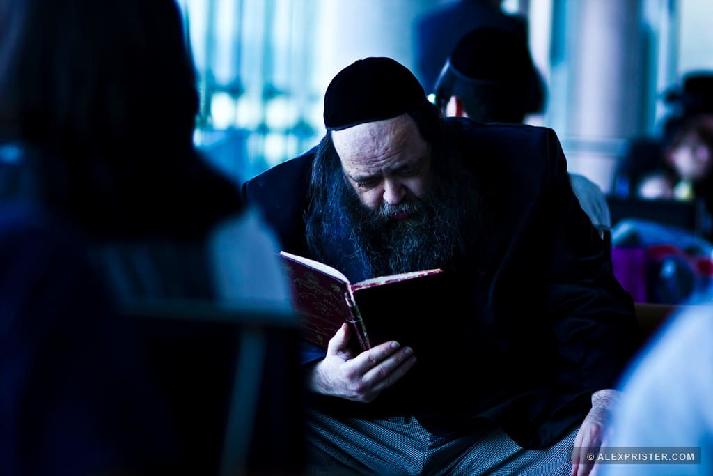 A bearded man in dark clothing reading a book in a softly blurred public setting