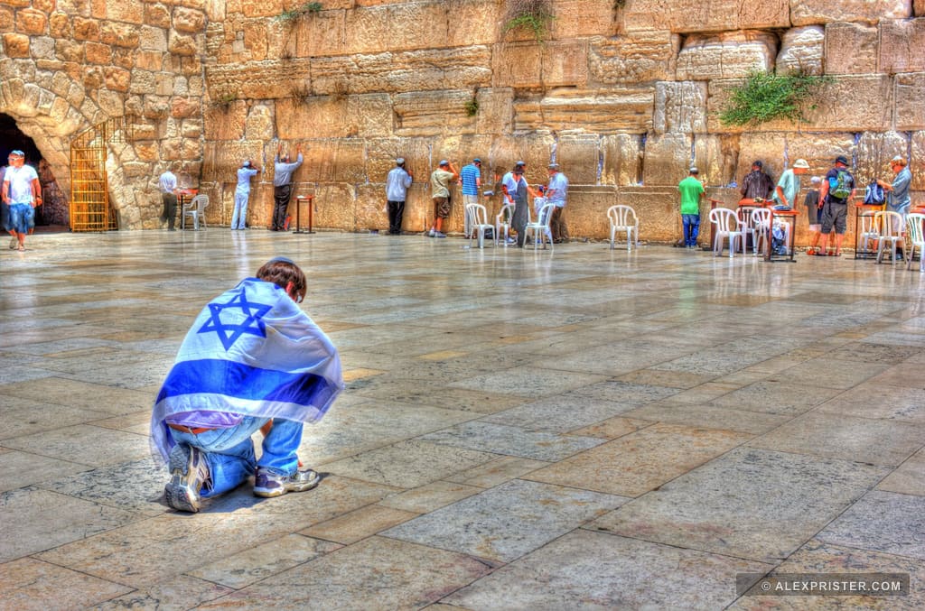 A person wrapped in a blue-and-white flag kneeling in a wet plaza before the Western Wall